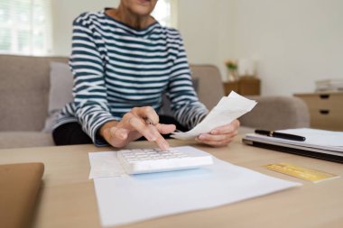 Finansal işler için hesap makinesi kullanan yaşlı bir kadın. Faturayı elinde tutuyor ve rahat bir oturma odasındaki ahşap bir masada oturuyor..