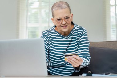 Bilgisayarda online alışveriş için kredi kartı kullanan yaşlı bir kadın modern bir evde rahatça oturuyor..