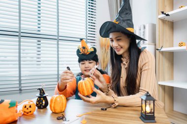 Cadı şapkası takan bir anne ve çocuğu bir masada balkabağı oyup cadılar bayramını aydınlık ve modern bir evde kutluyorlar..