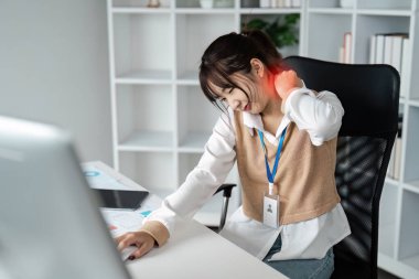 Modern bir ofiste masa başında oturan genç bir kadın, uzun süreli bilgisayar kullanımından dolayı rahatsızlığını ya da gerginliğini gösteren acılar içinde boynunu tutuyor..