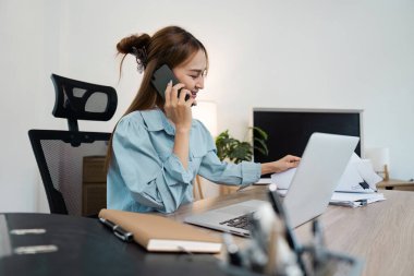 Masasında birden fazla iş yapan bir iş kadını, dizüstü bilgisayar kullanıyor ve modern ofis ortamında telefonla konuşuyor..