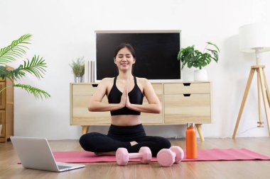 Evde yoga yapan genç bir kadın, halterler ve su şişesi gibi sağlıklı malzemelerle çevrili, modern oturma odasında..