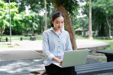 Parkta dizüstü bilgisayarda çalışan genç profesyonel bir kadın modern iş esnekliği ve uzaktan kumandalı çalışmalar sergiliyor..