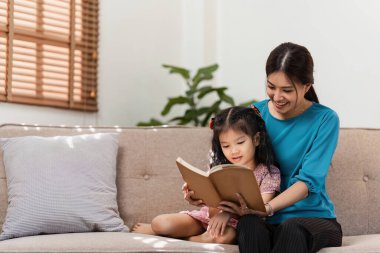 Sevgi dolu bir anne ve genç kızı, iyi aydınlatılmış, modern bir oturma odasında, rahat bir kanepede kitap okuyarak kaliteli zaman geçirmekten zevk alırlar..