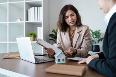 Emlakçı, modern bir ofisteki müşteriye emlak detaylarını açıklıyor. Model bir ev ve belgeler sergiliyor..