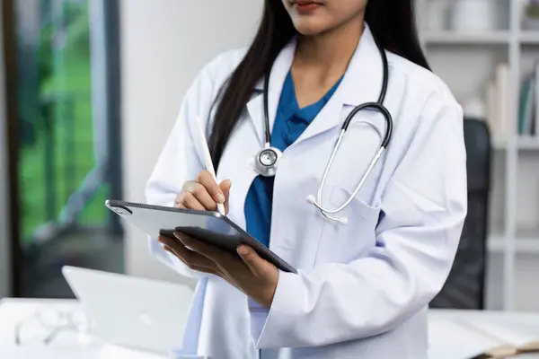 Modern bir muayenehanede dijital tablet kullanan beyaz önlüklü kadın doktor, sağlık hizmetlerindeki teknolojiyi vurguladı.