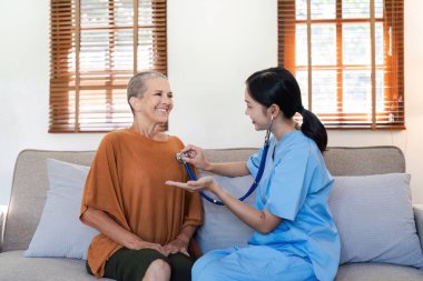 Mavi önlüklü bir hemşire, gülümseyen bir yaşlı kadının kalp atışlarını kontrol etmek için steteskop kullanıyor..