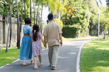 Üç kişilik bir aile, yemyeşil bir parkta asfalt bir yolda yürüyor güneşli bir günün tadını çıkarıyor ve birlikte kaliteli zaman geçiriyorlar..