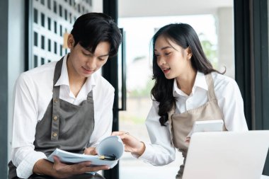 Önlüklü iki küçük işletme sahibi dijital tablet ve defter kullanarak modern bir kafede iş planlarını tartışıyor..