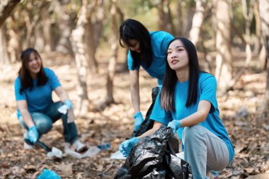 Mavi gömlekli ve eldivenli bir grup genç gönüllü, ormanda çöp topluyor, takım çalışması ve çevrenin korunmasına olan bağlılıklarını gösteriyorlar..