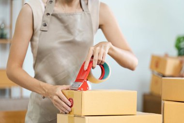 Küçük bir işletme sahibi karton bir kutuyu mühürlemek için bant kullanıyormuş. Ev ofis ortamında nakliye için ürünler hazırlıyormuş..