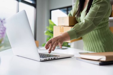 Küçük bir işletme sahibi, paketleme kutularını modern ofis ortamında tutarken çevrimiçi siparişleri yönetmek için dizüstü bilgisayar kullanır..