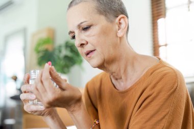 Elderly woman holding a pill and a glass of water, focusing on her health and wellness in a cozy home environment. clipart