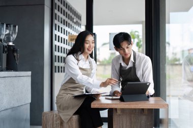 Önlüklü iki genç girişimci modern bir kafe ortamında dijital tablet kullanarak iş stratejisini tartışıyor, başlangıç kültürü ve takım çalışması sergiliyorlar..