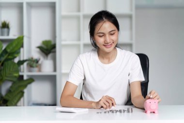 Beyaz gömlekli genç bir kadın ev ofisinde pembe bir kumbarayla para biriktiriyor. Finansal planlamayı ve bütçeyi sembolize ediyor..