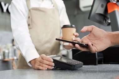 Önlüklü bir barista, kahve dükkanındaki akıllı telefonuyla müşteriye ücretsiz ödeme yapar..