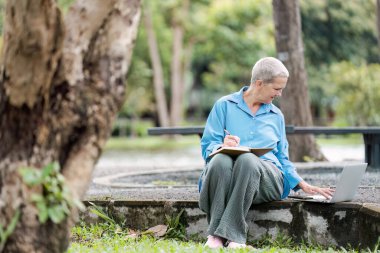 Yaşlı bir kadın parkta bankta oturuyor, dizüstü bilgisayar kullanıyor ve doğayla iç içe bir defter yazıyor..