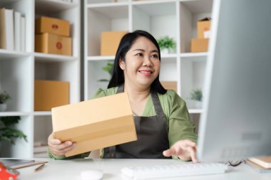 Elinde bir paket olan gülümseyen bir kadın ve arka planda raflar olan bir ofis bilgisayarında çalışıyor..