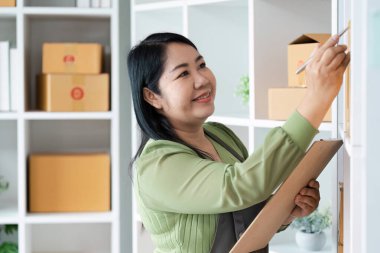 Modern bir ofis raflarında paketleri düzenleyen gülümseyen bir kadın, elinde bir pano ve kalem, düzgünce düzenlenmiş kutularla çevrili..