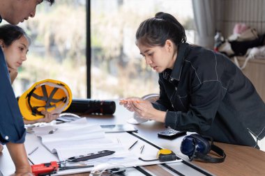 Siyah ceketli genç bayan mühendis inşaat projesinde ekiple birlikte çalışıyor, modern ofis ortamında planları ve aletleri inceliyor..