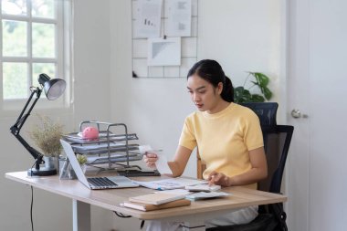 Sarı tişörtlü genç bir kadın ofis masasında oturuyor, bir dizüstü bilgisayar, belgeler ve hesap makinesiyle finansman yönetiyor..