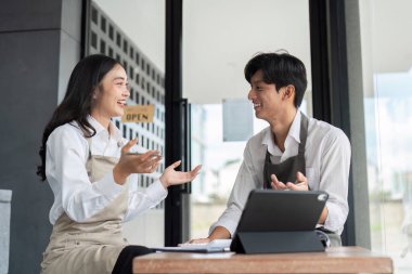Önlüklü iki genç Asyalı kafe çalışanı modern bir kahve dükkanında dijital tabletle iş planlarını tartışıyor..