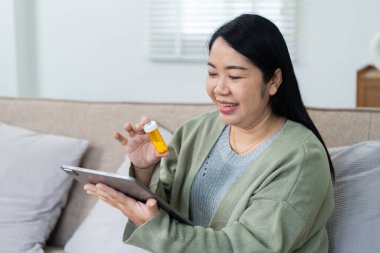 Elinde reçeteli bir şişeyle dijital tablet kullanan mutlu bir kadın evinde konforlu bir şekilde çevrimiçi tıbbi konsültasyona katılıyor..