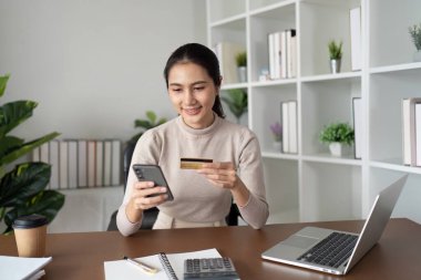 Modern bir ev ofisinde bilgisayar ve bitkilerle online alışveriş yapmak için akıllı telefon ve kredi kartı kullanan gülümseyen genç bir kadın..