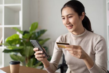Çevrimiçi alışveriş için akıllı telefon ve kredi kartı kullanan gülümseyen bir kadın yeşil bitkiler ve kahve fincanı olan modern bir ev ofisinde..