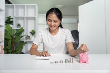 Beyaz tişörtlü, gülümseyen genç bir kadın bozuk paraları sayıyor ve modern bir ofis masasındaki domuz kumbarasına koyuyor..