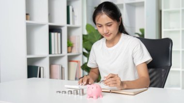 Beyaz gömlekli genç bir kadın, kumbarası ve ofis masasında madeni para tasarrufu yaparak mali planlama ve bütçeye odaklanıyor..