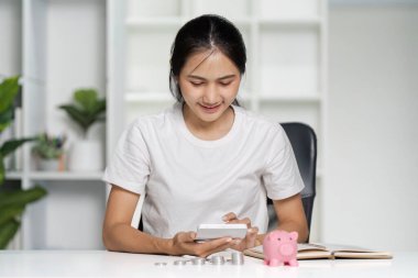 Genç bir kadın, banka ve not defteriyle mali işleri yürütmek için akıllı telefonunu kullanıyor. Tasarruf ve bütçeyi sembolize ediyor..