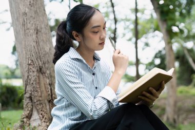 Parktaki çimlerin üzerinde oturan genç bir kadın, kitap okuyor ve gülümsüyor, ağaçlar ve yeşilliklerle çevrili..