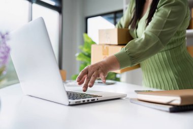 Küçük bir işletme sahibi, paketleme kutularını modern ofis ortamında tutarken çevrimiçi siparişleri yönetmek için dizüstü bilgisayar kullanır..