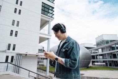 Kulaklık takan ve kot ceket giyen genç bir adam modern şehir ortamında akıllı telefonunu ve modern mimariyi kullanıyor..