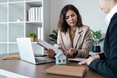 Emlakçı, modern bir ofisteki müşteriye emlak detaylarını açıklıyor. Model bir ev ve belgeler sergiliyor..