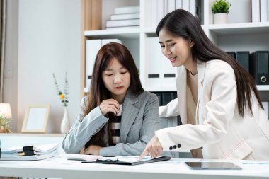 İş kıyafeti giymiş iki kadın modern ofis ortamında bir masada belgeleri tartışıyor, takım çalışması ve işbirliği sergiliyorlar..