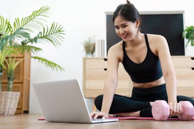 Bir kadın, rehberlik için dizüstü bilgisayar ve kuvvet eğitimi için halterler kullanarak, modern bir oturma odasında refahı sağlamak için ev antrenmanı yapar..