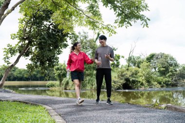 Parkta yürüyen ve sohbet eden genç bir çift, ellerinde su şişeleri, güneşli bir günde yürüyüş sonrası eğleniyorlar..