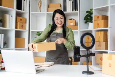 Küçük bir işletme sahibi, modern e-ticaret çalışma alanını dizüstü bilgisayar ve halka ışığı ile göstererek, bir ev ofisinde sevkiyat için paketler hazırlar..