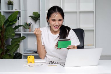 Neşeli bir kadın, elinde bir kredi kartı tutarken ve modern bir ofis ortamında dizüstü bilgisayar kullanırken tasarruf başarısını kutluyor..