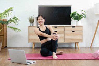 Atletik bir kadın modern bir oturma odasında pembe bir paspasla yoga yaparken dizüstü bilgisayar kullanıyor..