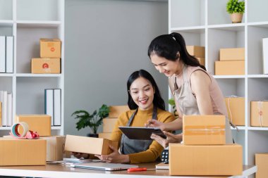 İki kadın küçük bir iş yürütüyor, nakliye kutuları düzenliyor ve modern ofis ortamında dijital tablet kullanıyorlar..