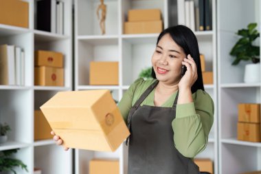 Önlüklü küçük bir işletme sahibi, modern bir ofiste raflar ve kutularla konuşurken siparişleri ve envanteri yönetiyor..