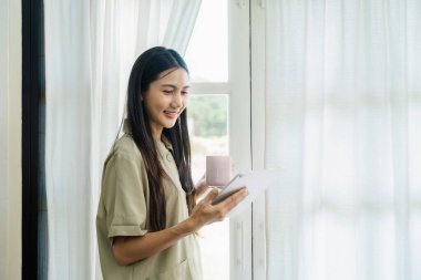 Genç bir kadın, pencerenin kenarında tablet kullanırken bir fincan kahvenin keyfini çıkarıyor. Modern teknoloji ve rahat bir yaşam tarzı sergiliyor..