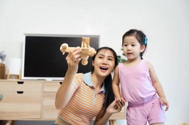 Neşeli bir anne ve genç kızı, parlak, modern bir oturma odasında tahta bir oyuncak uçakla oynuyorlar..