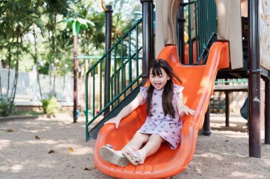 Mutlu çocuk, yeşil ve güneş ışığıyla çevrili oyun parkında eğlenceli bir kaydırağın keyfini çıkarıyor..