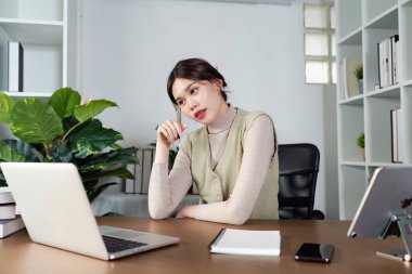 Evden çalışan genç bir kadın, dizüstü bilgisayarı ve not defteriyle bir masada oturuyor, modern bir ofis ortamında..