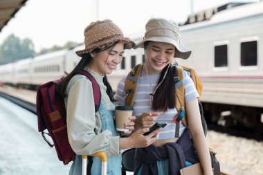 Tren istasyonunda sırt çantalı ve şapkalı iki genç kadın gülümsüyor ve telefona bakıyor. Seyahat maceralarına hazırlanıyorlar..