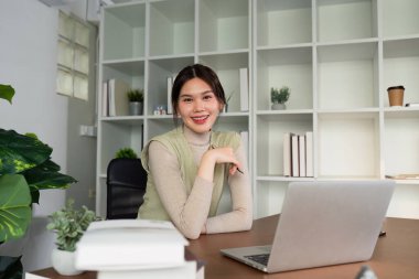 Modern ofis ortamında masa başında çalışan kendine güvenen genç bir kadın kameraya dizüstü bilgisayarla gülümsüyor ve arka planda raflar düzenliyor..